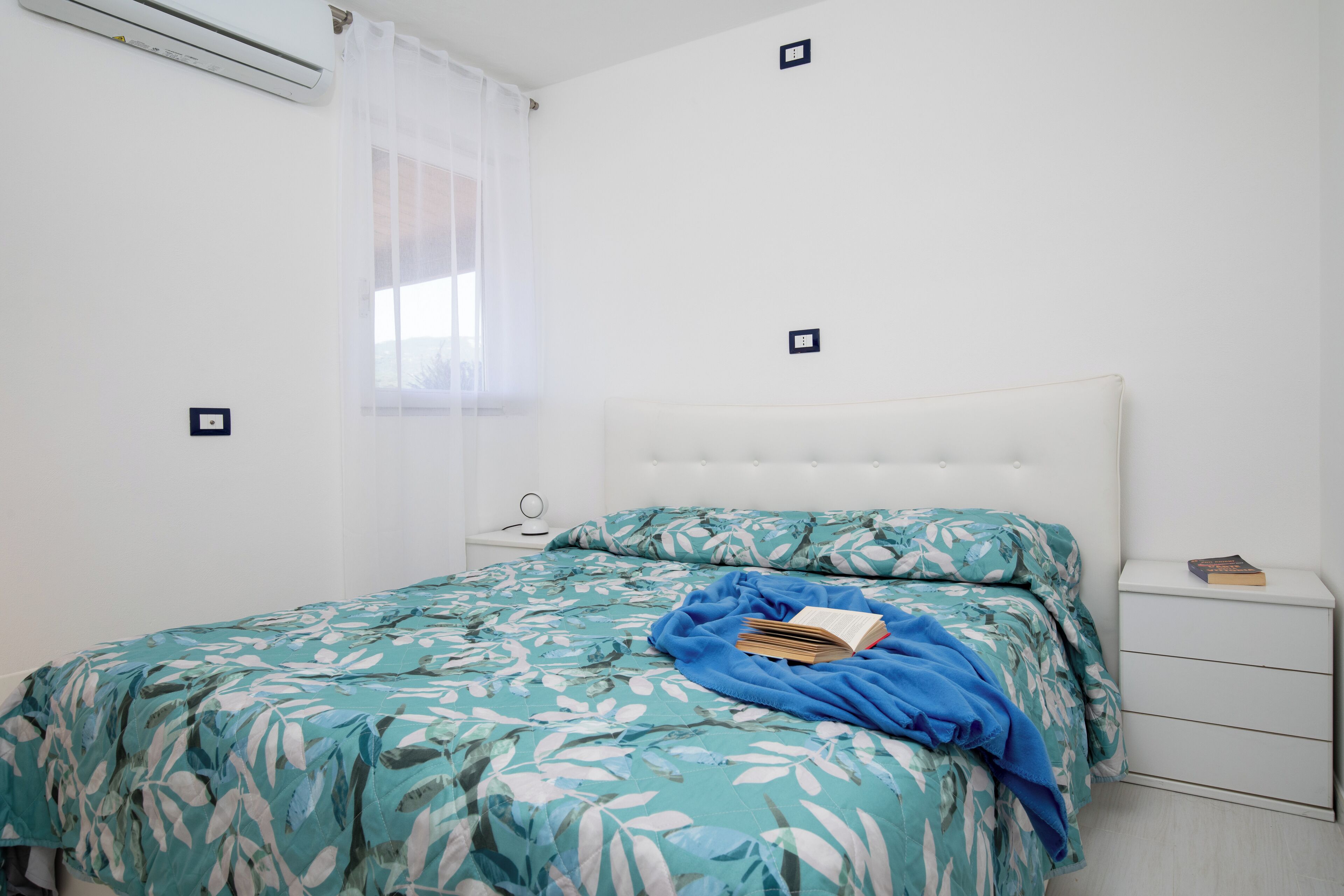 Ein Schlafzimmer mit einem Bett, einem Nachttisch, einem Fenster mit Vorhängen und einer Wandklimaanlage.