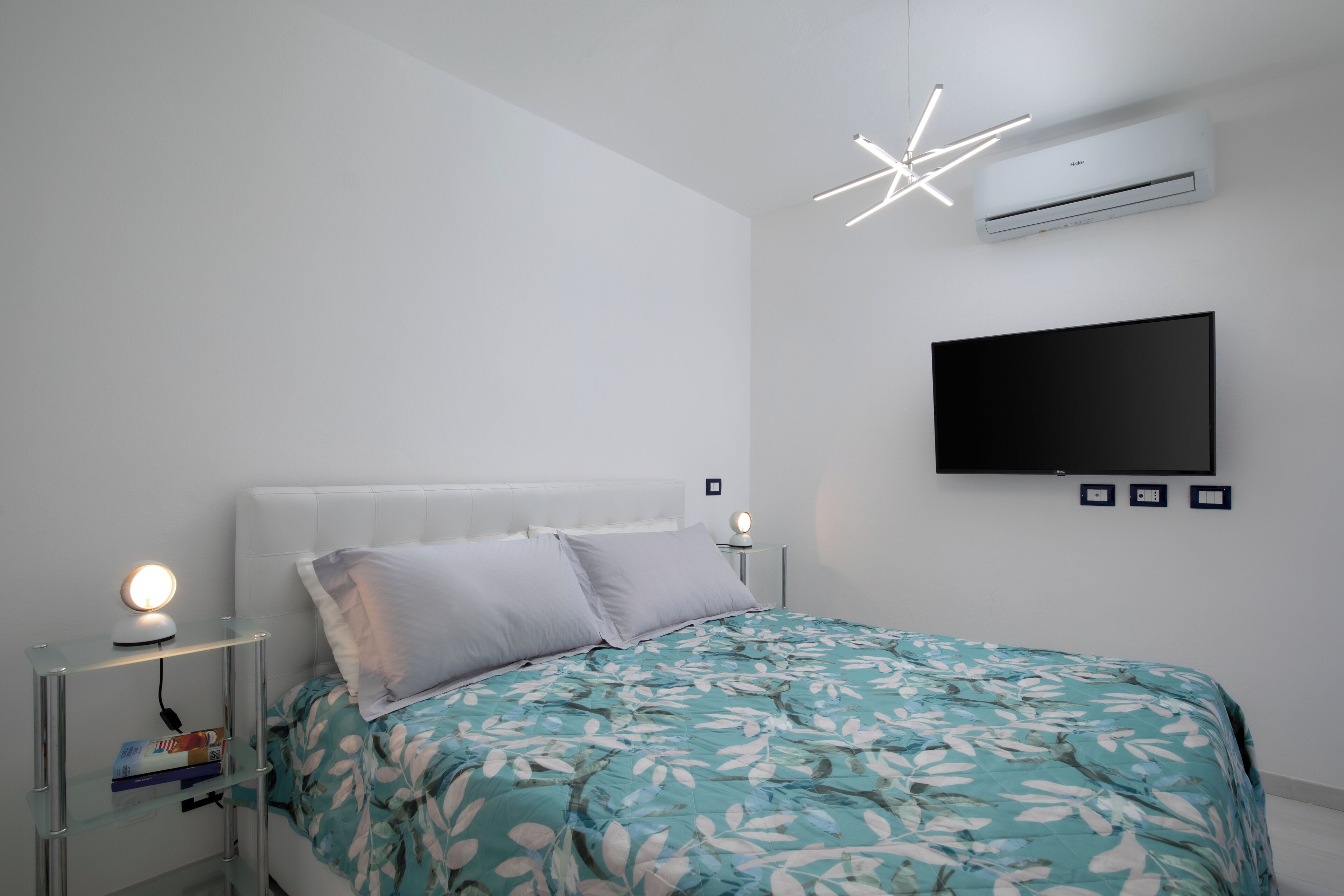 A bedroom with a bed, a bedside table with a lamp, a wall-mounted TV, and an air conditioner.