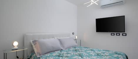 A bedroom with a bed, a bedside table with a lamp, a wall-mounted TV, and an air conditioner.