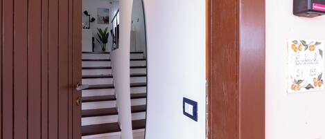 A hallway with a wooden door, a staircase, a wall-mounted mirror, and a sign with fruit illustrations.