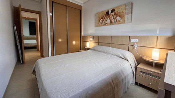 A hotel room with a bed, bedside table, nightstand, and a painting on the wall.