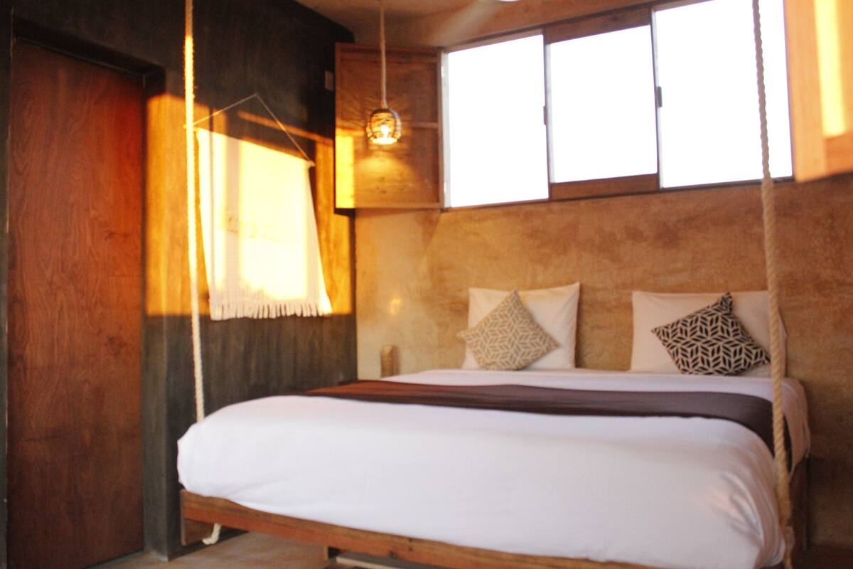 A wooden-framed bed with white bedding and patterned pillows, a hanging light fixture, and a window with a view.