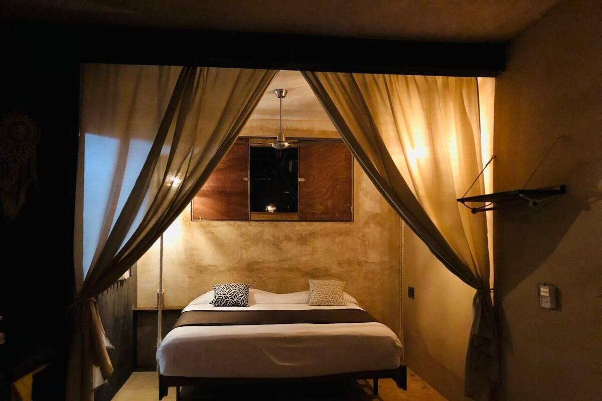 A bedroom with a bed, curtains, a hanging light fixture, and a small shelf above the bed.