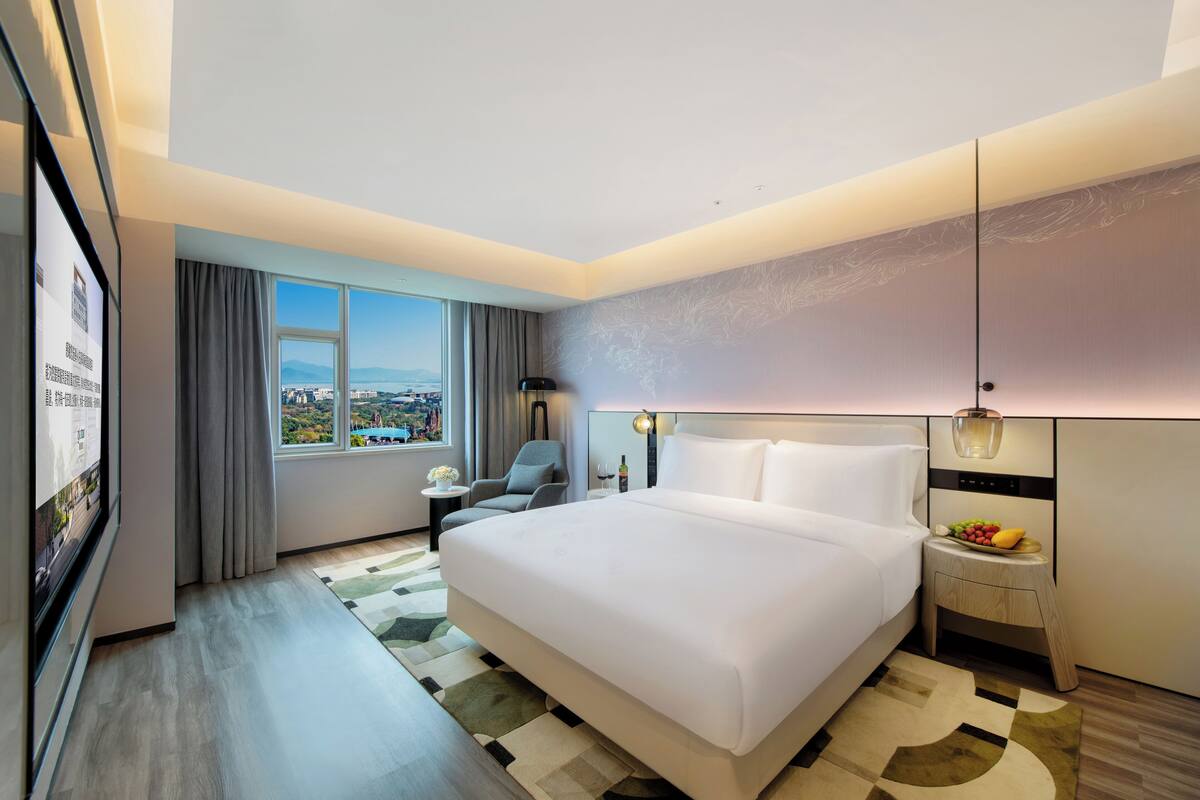 A modern hotel room with a large bed, a desk, a chair, a nightstand with a fruit bowl, a lamp, and a large window with a city view.