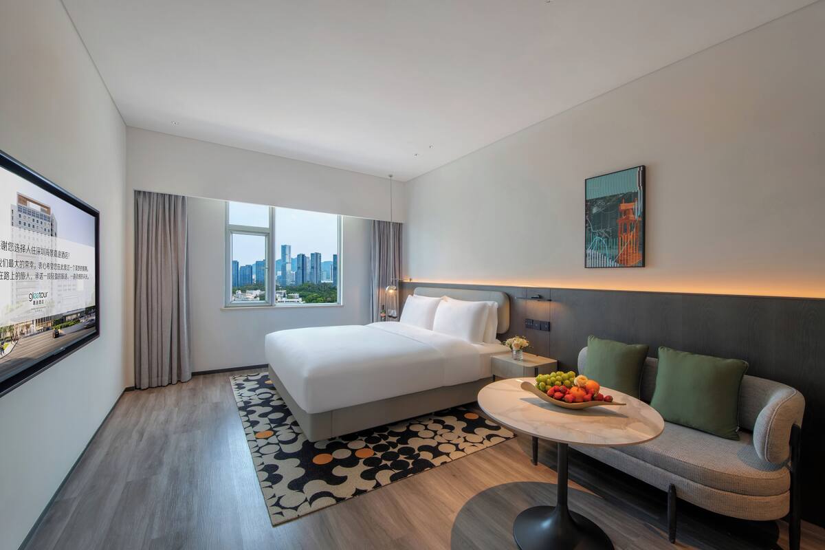 A modern hotel room with a large bed, a sofa, a dining table with fruit, and a city view.