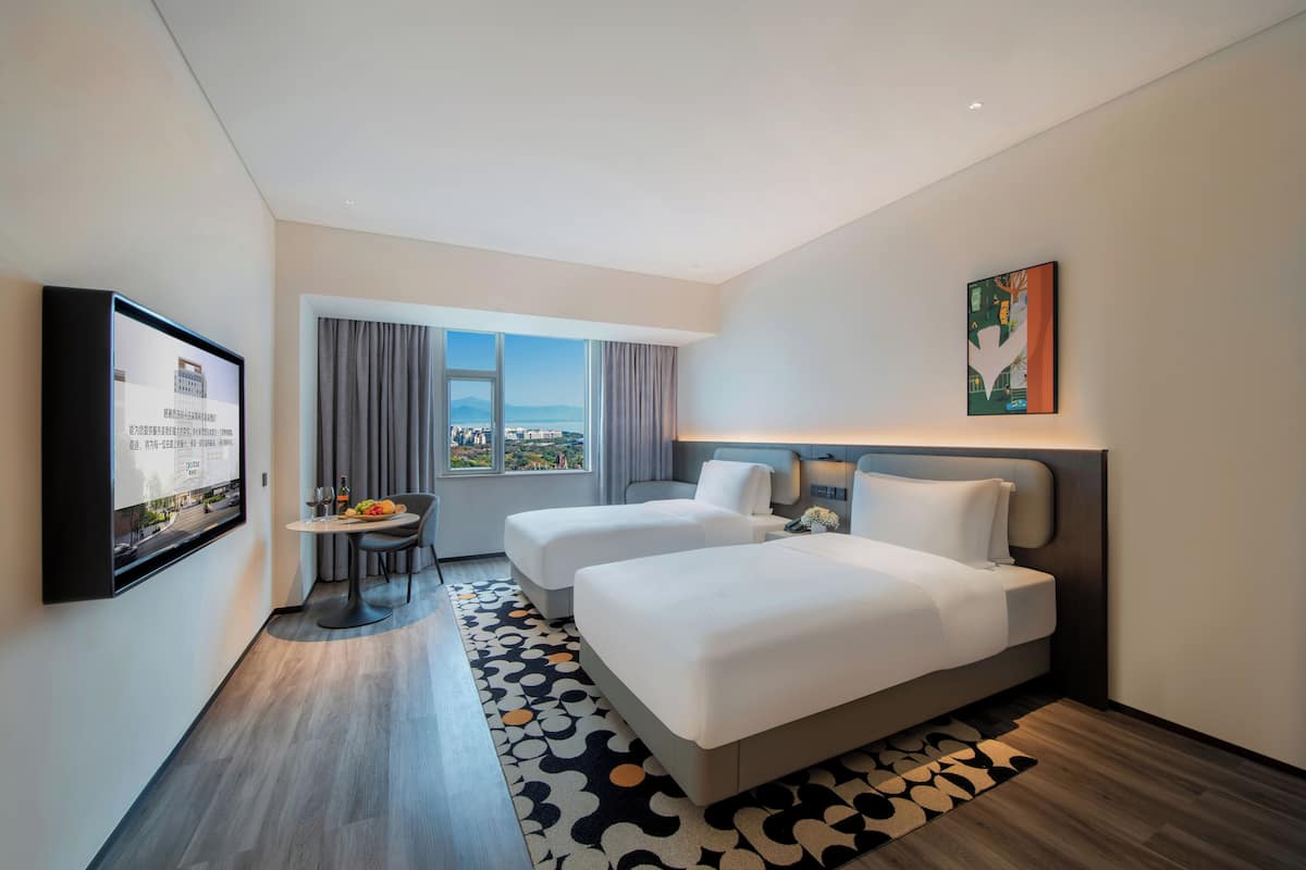 A hotel room with two beds, a dining table with chairs, a TV mounted on the wall, and a large window with a view.