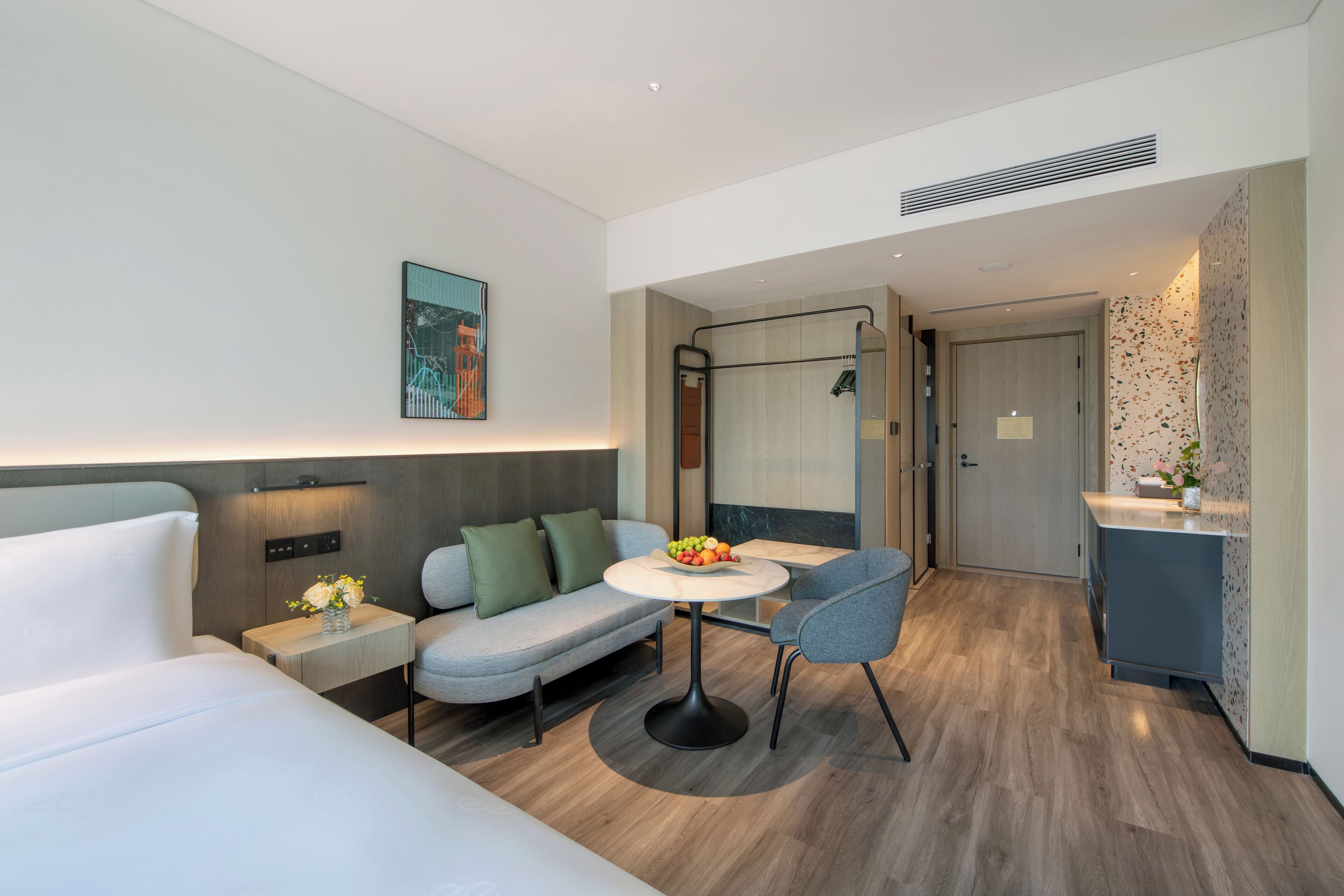 A modern hotel room with a bed, a sofa, a round table with a fruit bowl, a small side table with flowers, and a bathroom visible through a glass door.