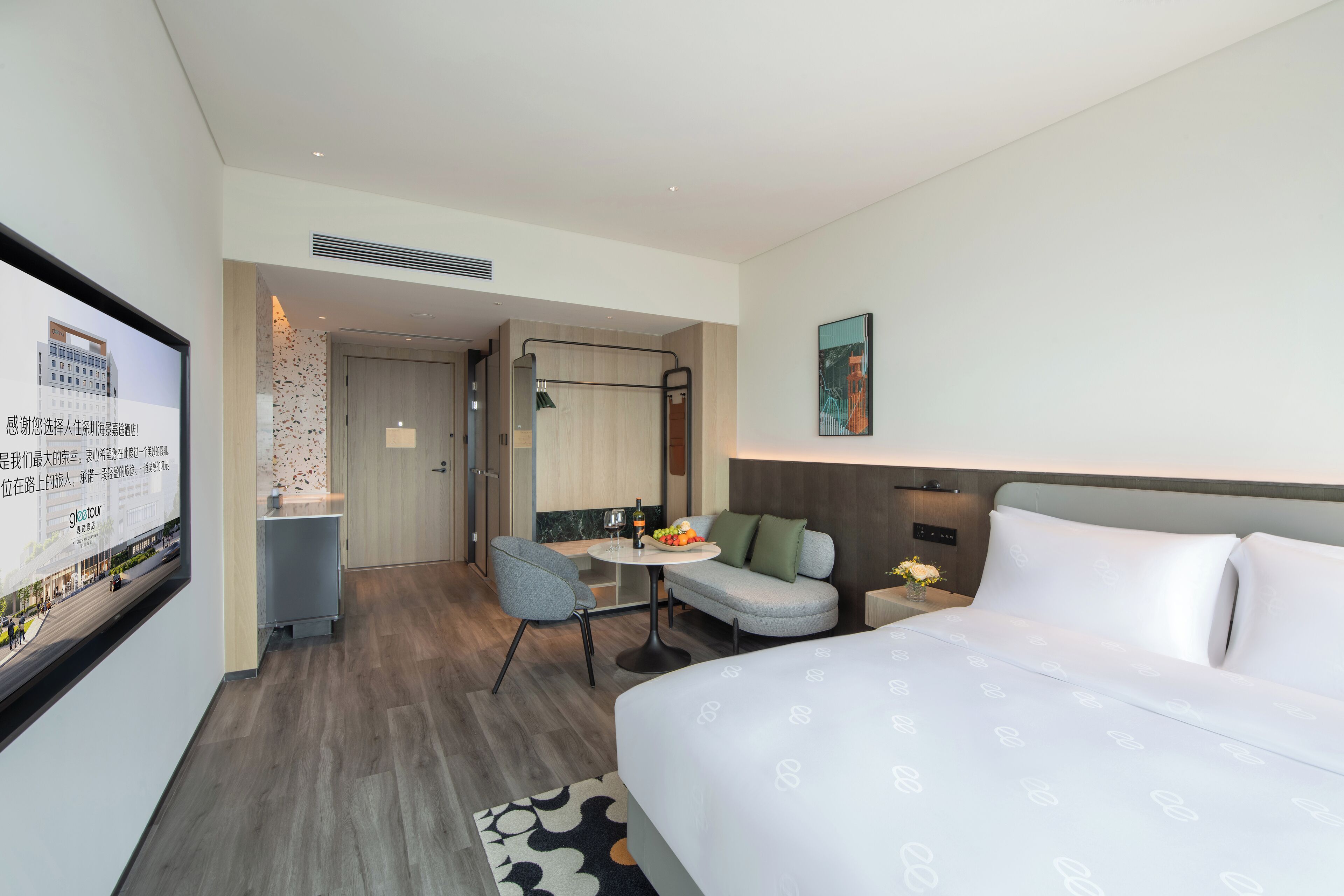 A modern hotel room with a large bed, a desk with a chair, a small table with a bowl of fruit, and a TV mounted on the wall.