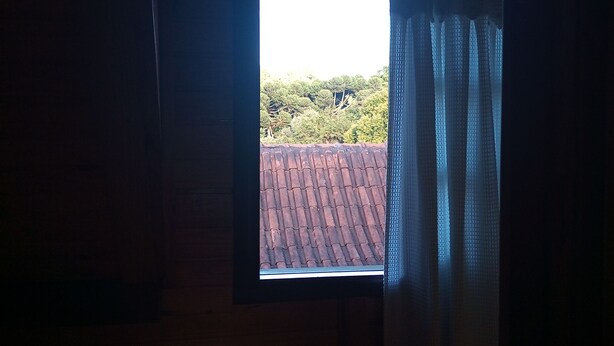 A small window with a view of a red-tiled roof and greenery outside. - Casa da Dine (Campos do Jordão)