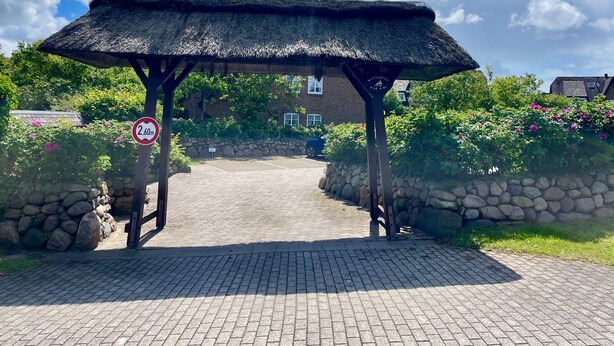 A thatched-roof entrance to a building with a paved pathway leading to it, surrounded by greenery and a stone wall.