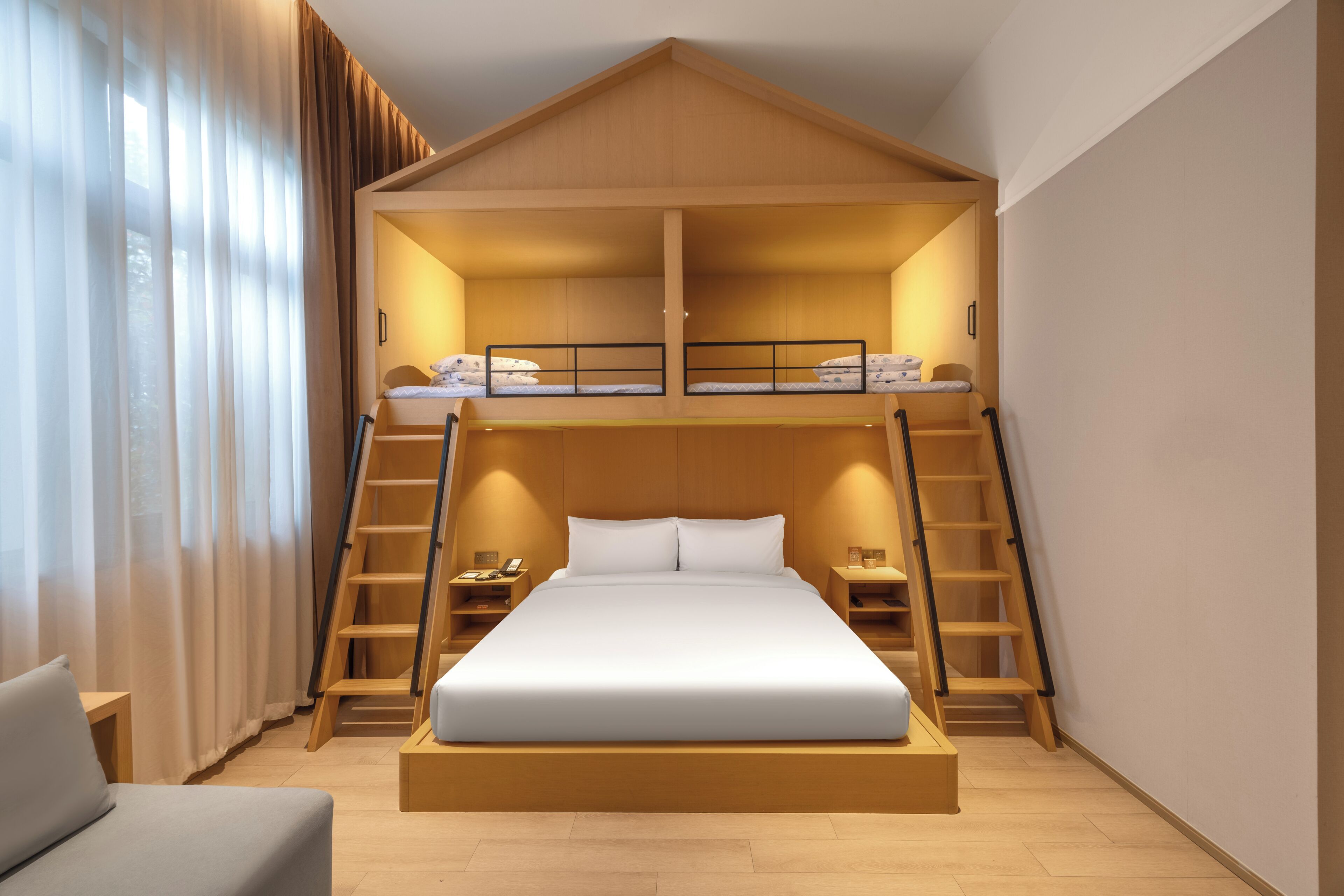 A modern hotel room with a bunk bed, wooden ladder, bedside tables, and a large bed.