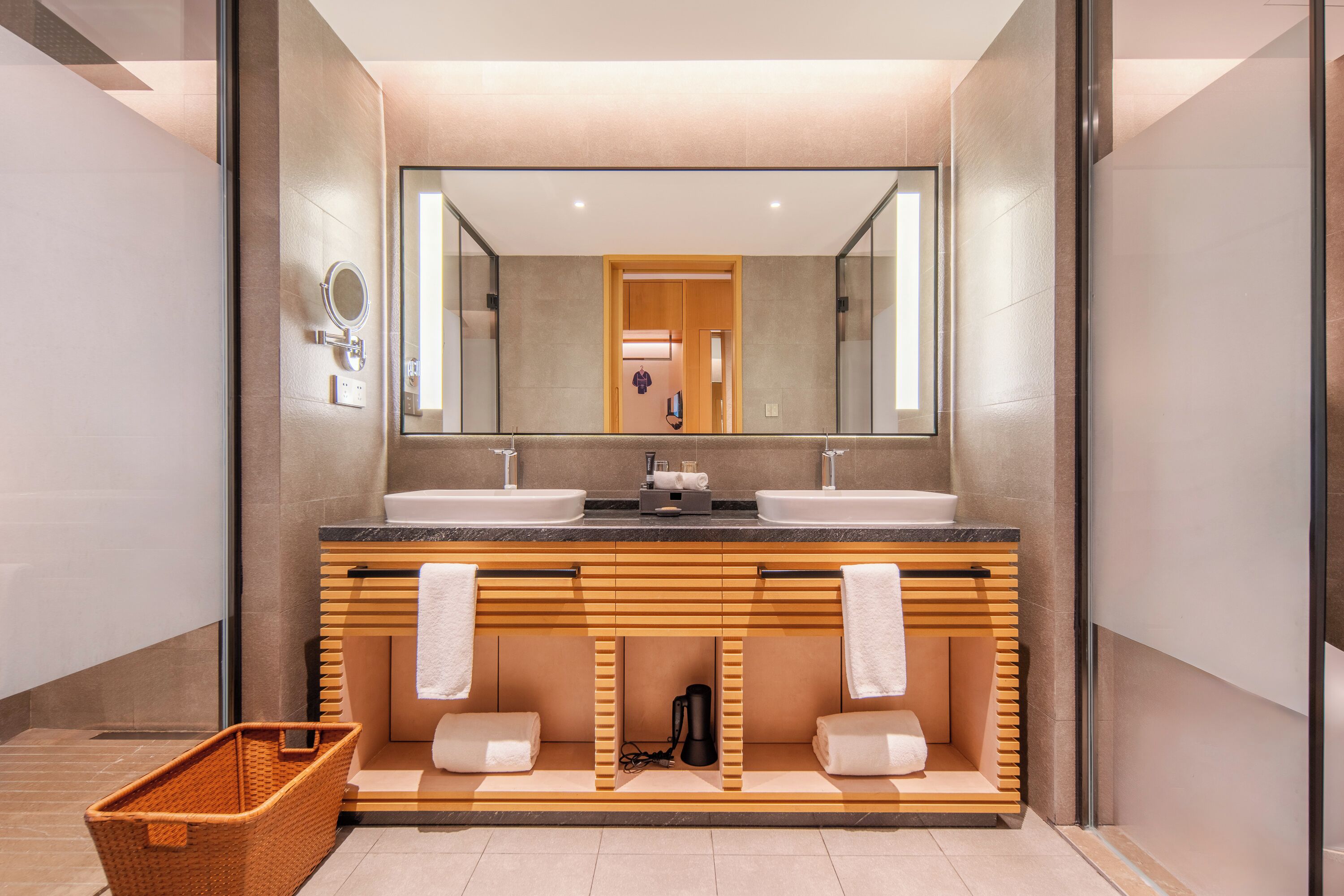 A modern bathroom with two sinks, a large mirror, wooden vanity, and a basket.