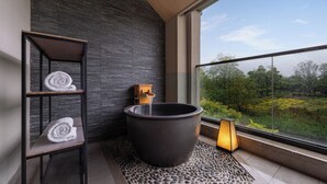 A modern bathroom with a large bathtub, a wooden shelf with towels, a stone-textured floor, and a view of greenery through a large window. - Hyakumangoku Onsen Hotel - Free Shuttle Service to Shanghai Disneyland (Shanghai)