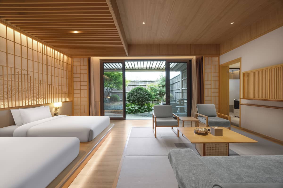 Une chambre d’hôtel moderne avec un plafond en bois, de grandes portes-fenêtres coulissantes donnant sur un jardin, un canapé, deux fauteuils et une petite table.