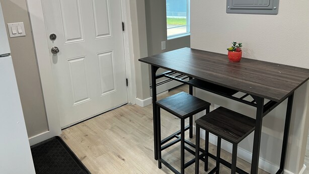 A small kitchen with a white door, a window, a wooden table with two stools, and a potted plant on the table.