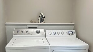 A laundry area with a front-loading washer and dryer, a wall-mounted ironing board, and a vent on the right.