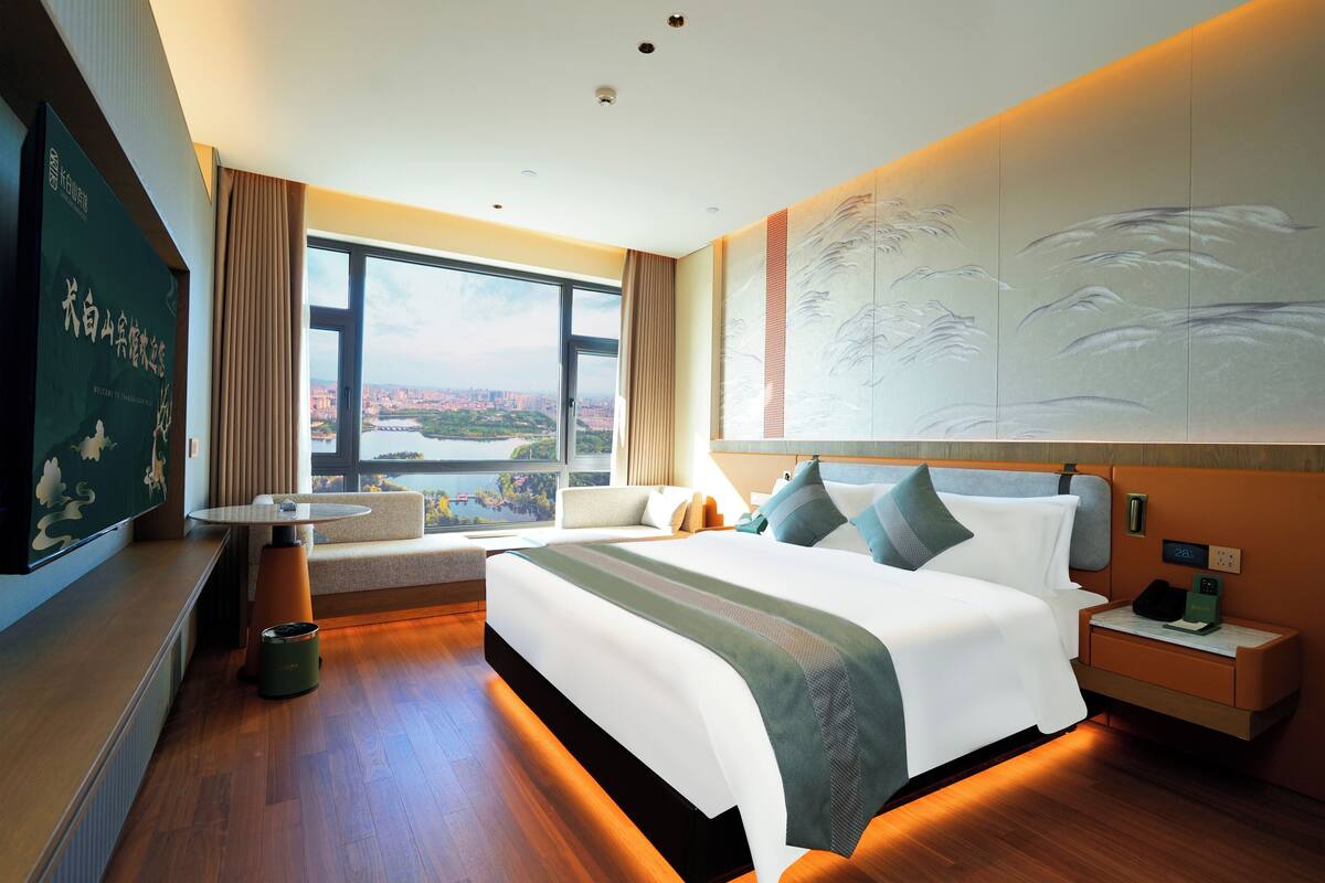 A modern hotel room with a large bed, a small table, a sofa, and a large window offering a city view.