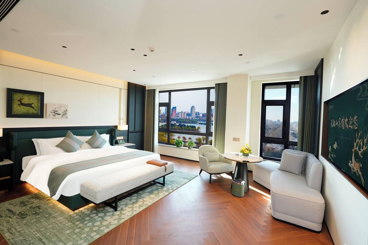 A modern hotel room with a large bed, a seating area, and a view of the cityscape.