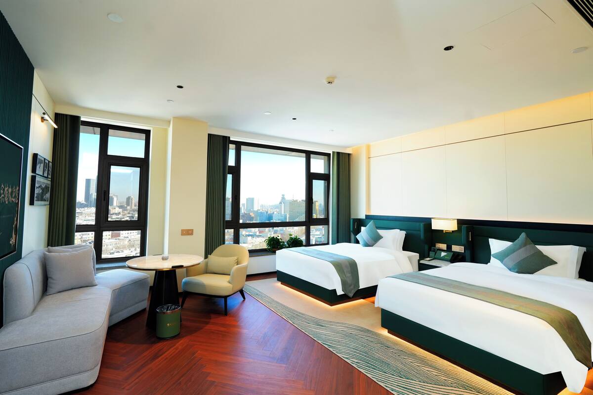 A hotel room with a large bed, a sofa, a small table, and a view of the city through large windows.