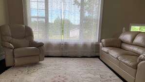 A living room with a beige armchair and sofa, a patterned rug, and a window with floral curtains. - Central PEI location minutes to golf and beaches.  (Cornwall)