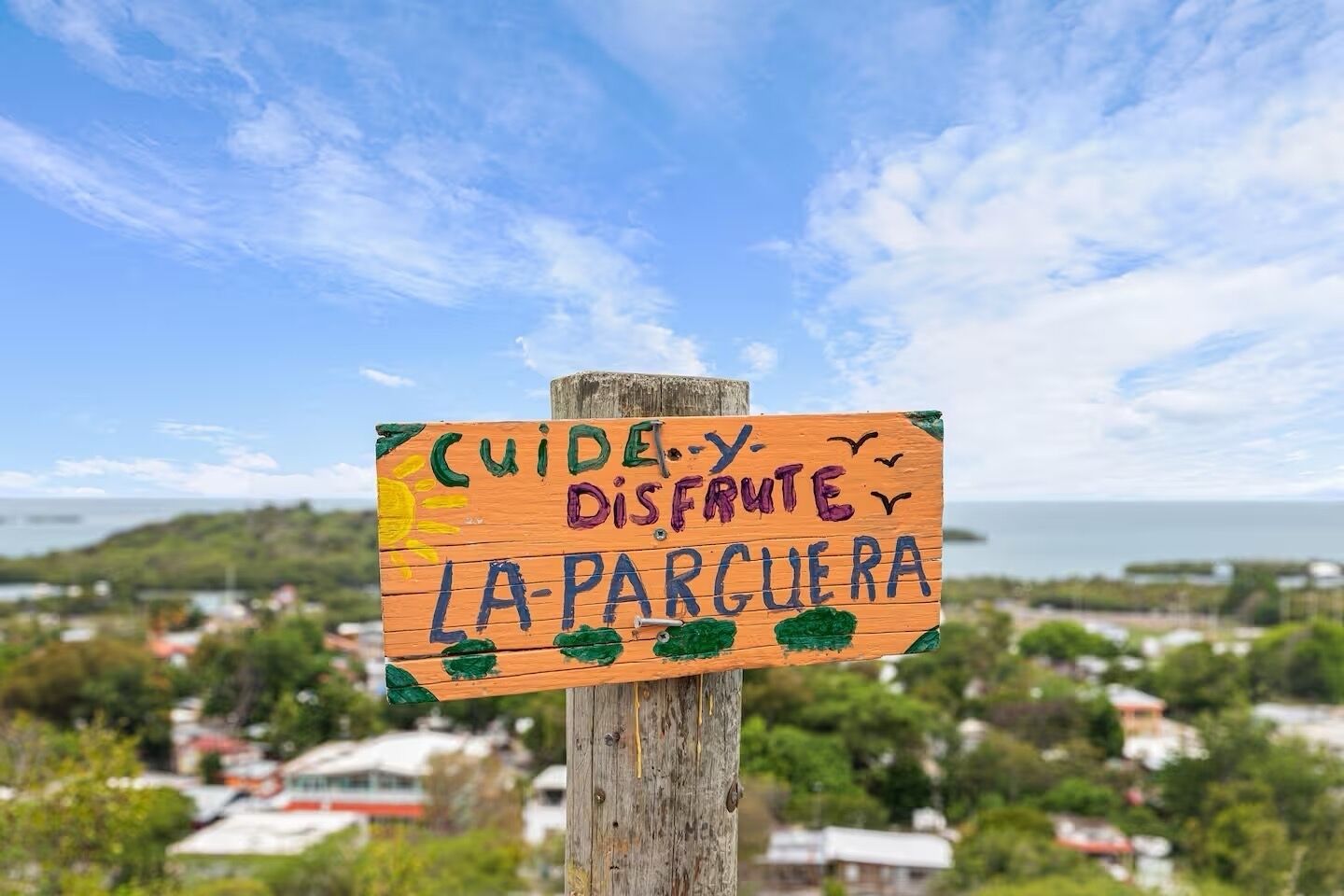 Un letrero de madera con texto y dibujos de colores, sujeto a un poste, con una vista panorámica de un pueblo costero y el océano al fondo.