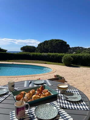 A poolside table with croissants and coffee, overlooking a pool and a scenic view of trees and hills.