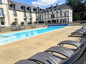 Piscina com espreguiçadeiras, cerca de madeira e um edifício com várias janelas e varandas.