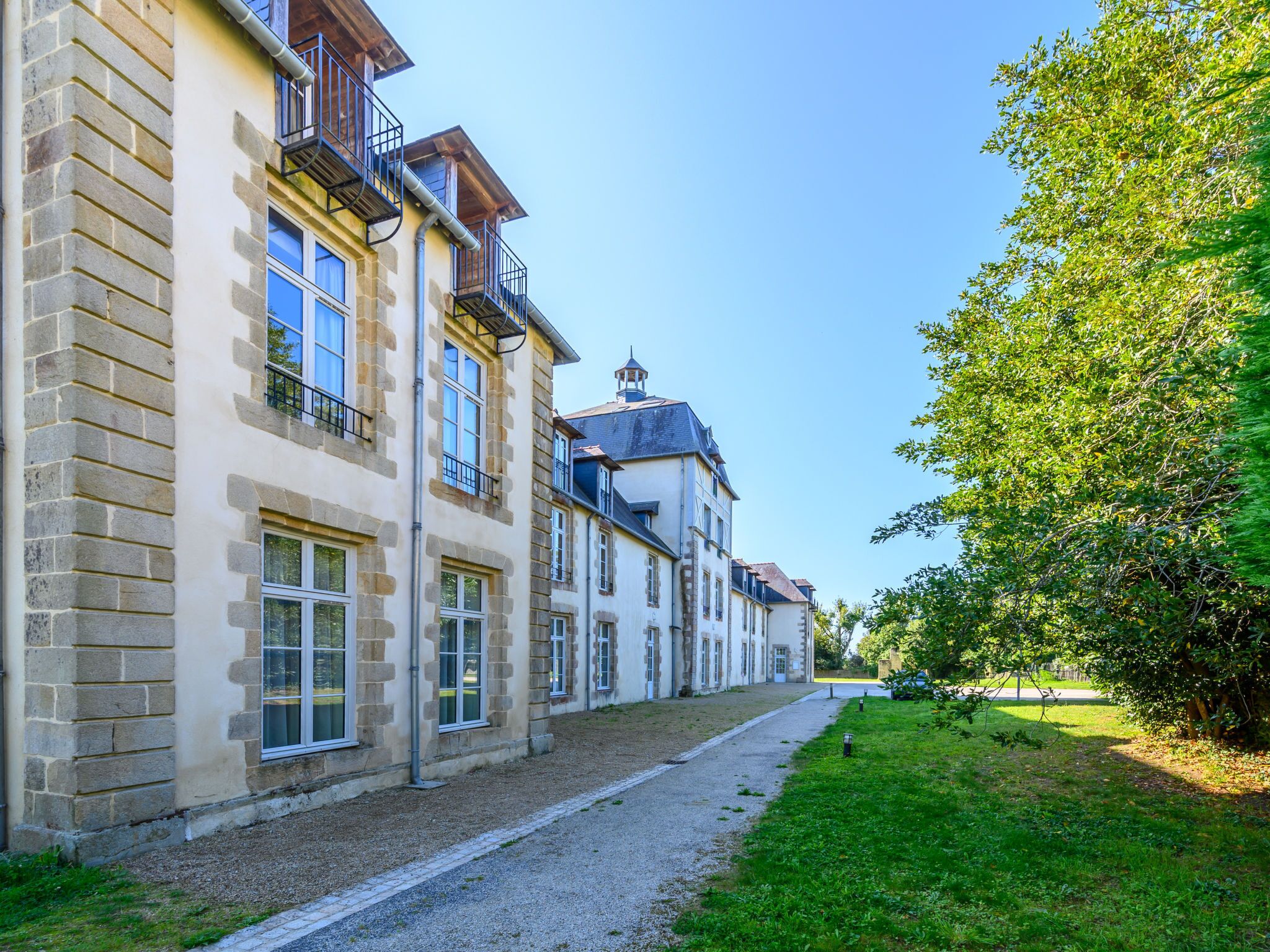 A row of stone buildings with multiple windows and balconies, a gravel path, and a grassy area with trees.