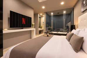 Ein modernes Hotelzimmer mit einem großen Bett, einem an der Wand befestigten Fernseher, einem Badezimmer mit Glasabtrennung und Blick auf die Stadtlandschaft.