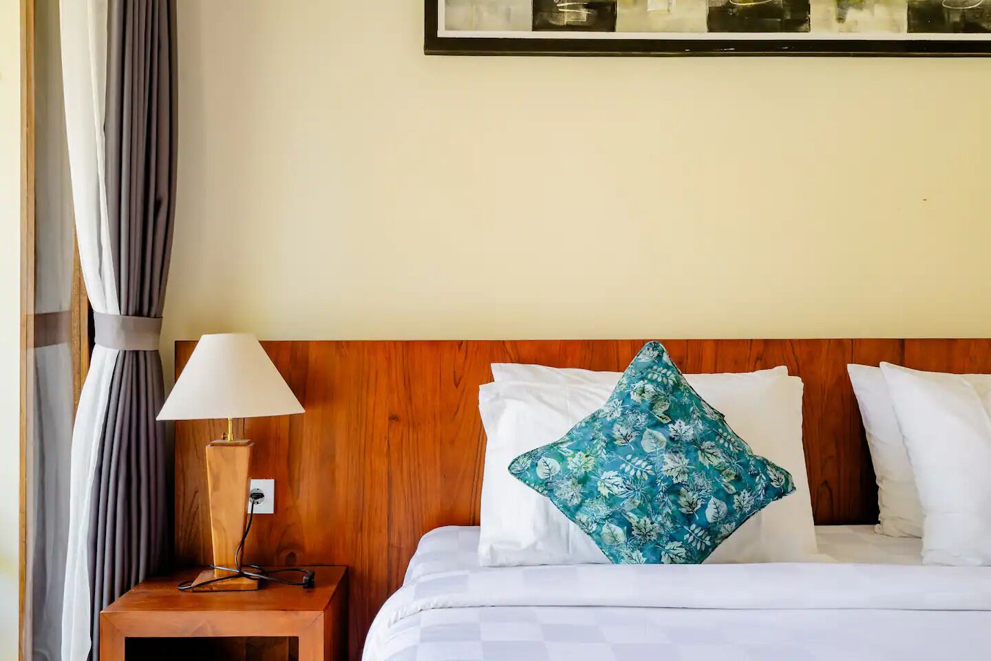 A hotel room with a wooden headboard, a bed with white bedding and a blue floral pillow, a bedside lamp, and a curtain on the left.