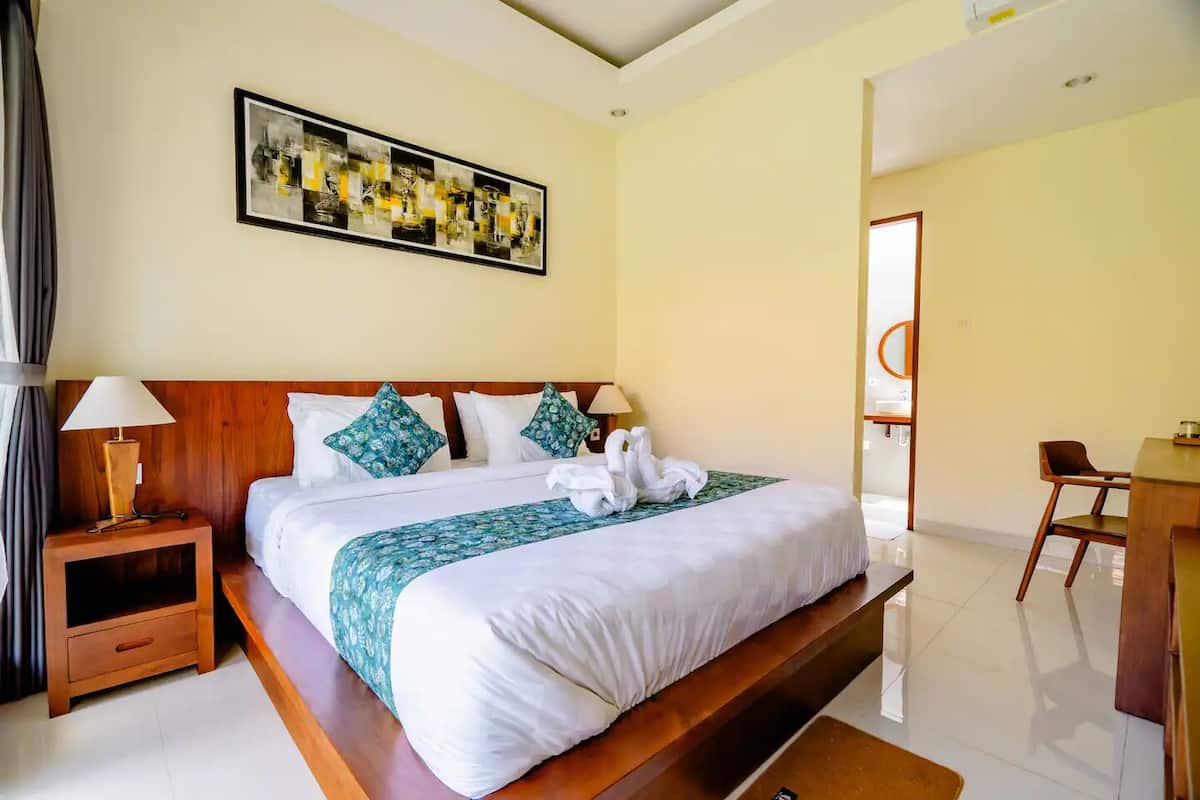 Une chambre d’hôtel avec un grand lit, une tête de lit en bois, une table de chevet avec une lampe, un bureau avec une chaise et un tableau au mur.