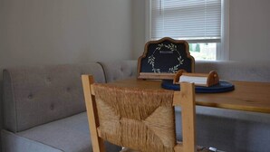 Une pièce avec un canapé gris, une table en bois, une chaise en osier, un tableau noir sur lequel est écrit le mot « BIENVENUE », et une fenêtre avec des stores.