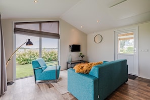 A modern living room with a sofa, a chair, a TV, and a large window offering a view of greenery. - Giethoorn Lakehouse Retreat (Giethoorn)