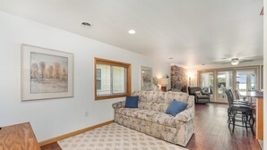 A spacious living room with a patterned sofa, a wooden coffee table, and a large window with blinds. - Higgins Lake Private Beachfront Home w/Dock, Boat Mooring, Sunset & Sunrise View (Roscommon)