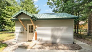 A small, single-story house with a green roof, a front door, and a welcome sign. - Higgins Lake Private Beachfront Home w/Dock, Boat Mooring, Sunset & Sunrise View (Roscommon)