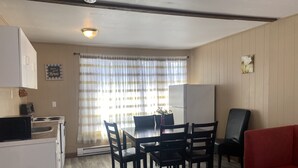 A compact kitchen with a microwave, stove, and refrigerator. There is a dining table with chairs and a red sofa. A window with curtains allows natural light into the room.