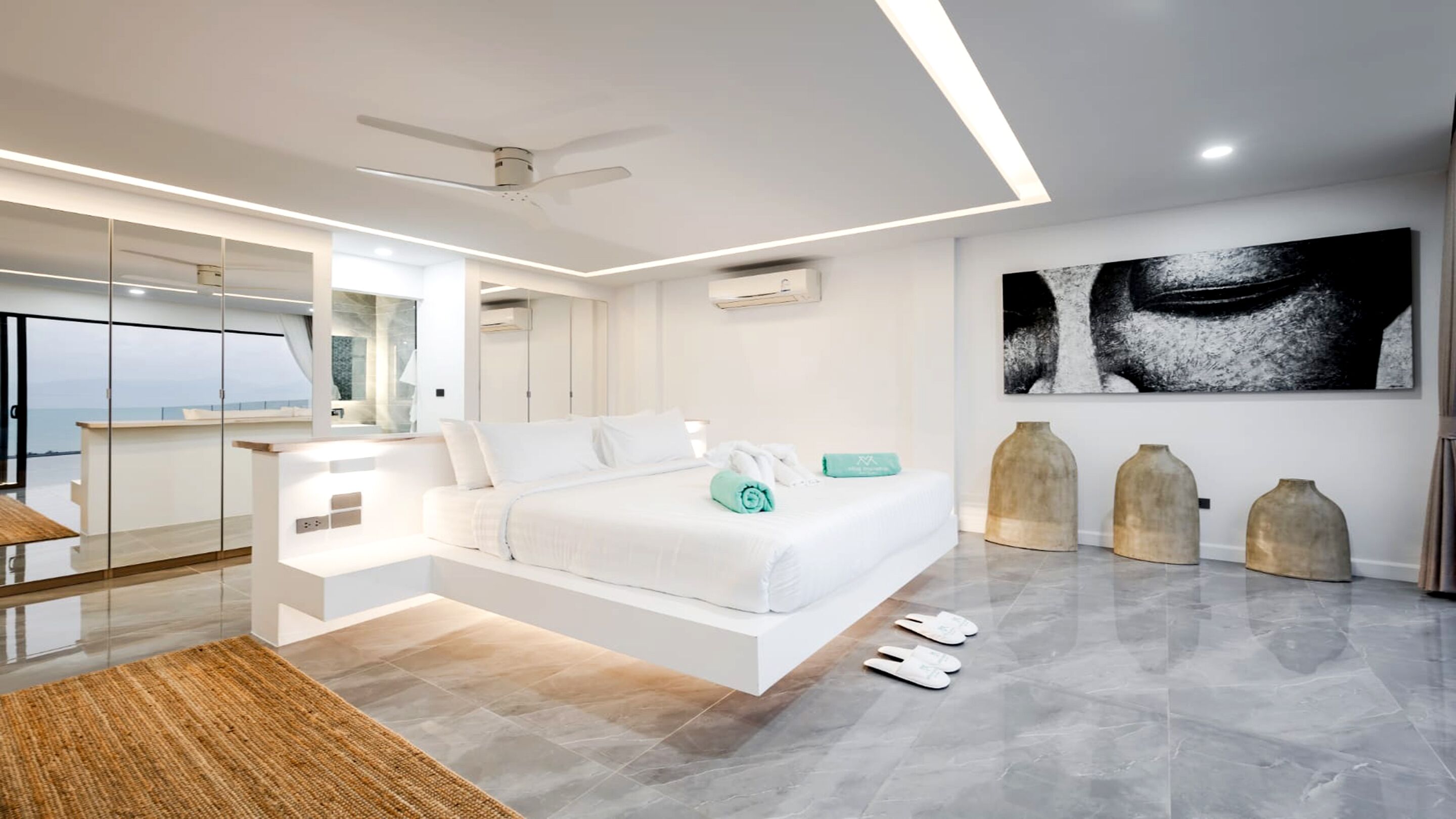 A modern bedroom with a bed, a large mirror, a ceiling fan, and a view of the sea.