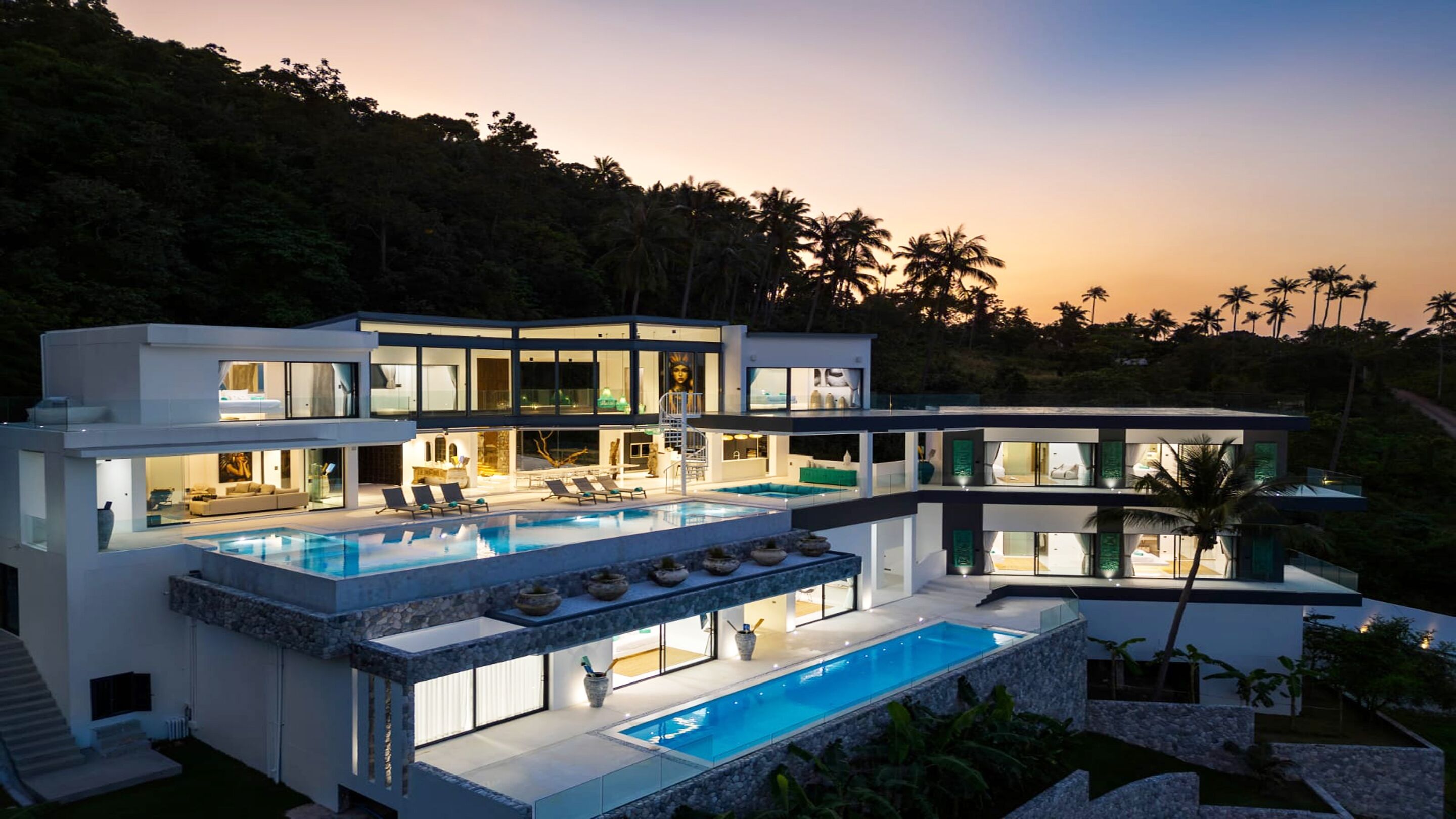 A modern multi-story house with a large swimming pool, multiple balconies, and a lush surrounding landscape.