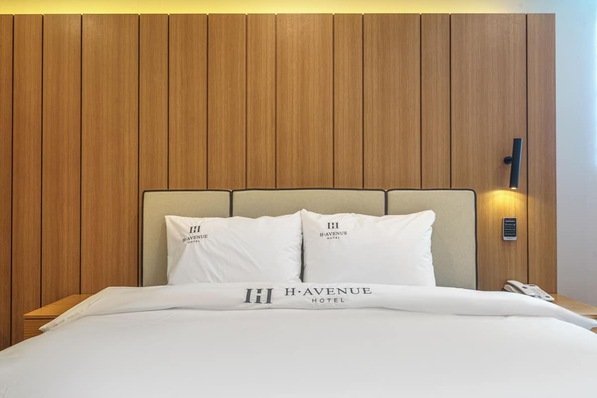 A hotel room with a wooden headboard, two pillows with the hotel logo, a bed with a white comforter, and a bedside phone.