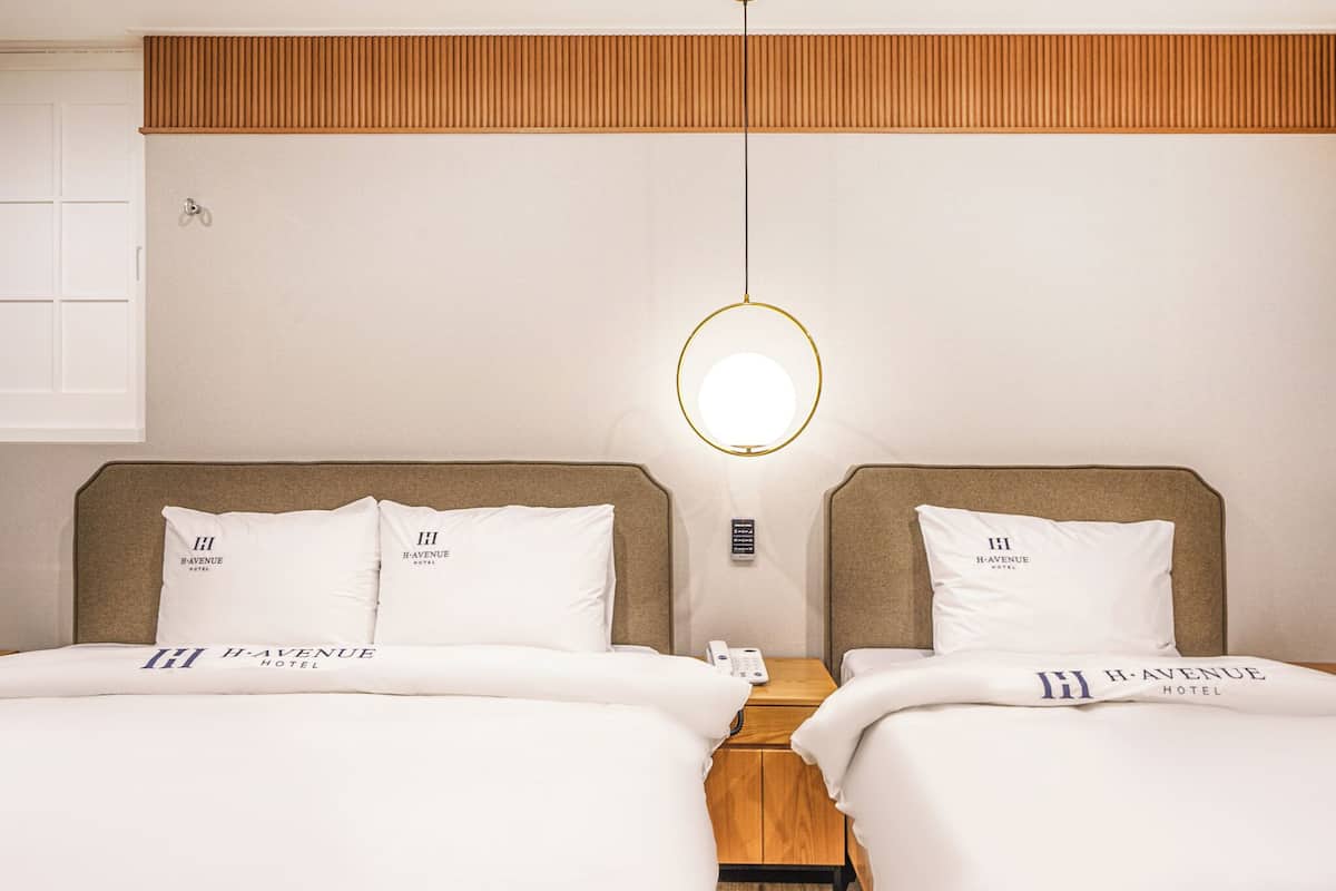 A hotel room with two beds, each with a headboard and pillows displaying the hotel logo. A bedside table with a phone and a lamp is visible.