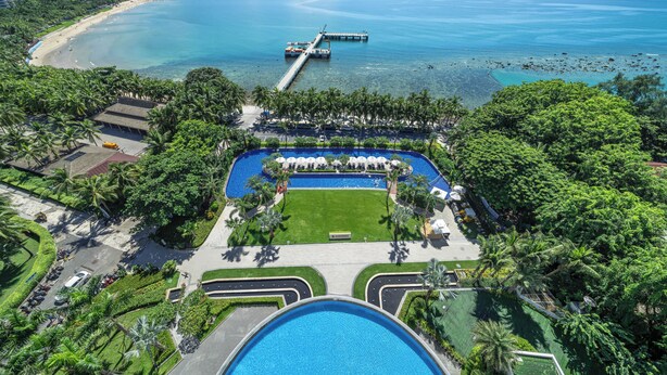 An aerial view of a resort with a large swimming pool, a beach, and a pier extending into the ocean.