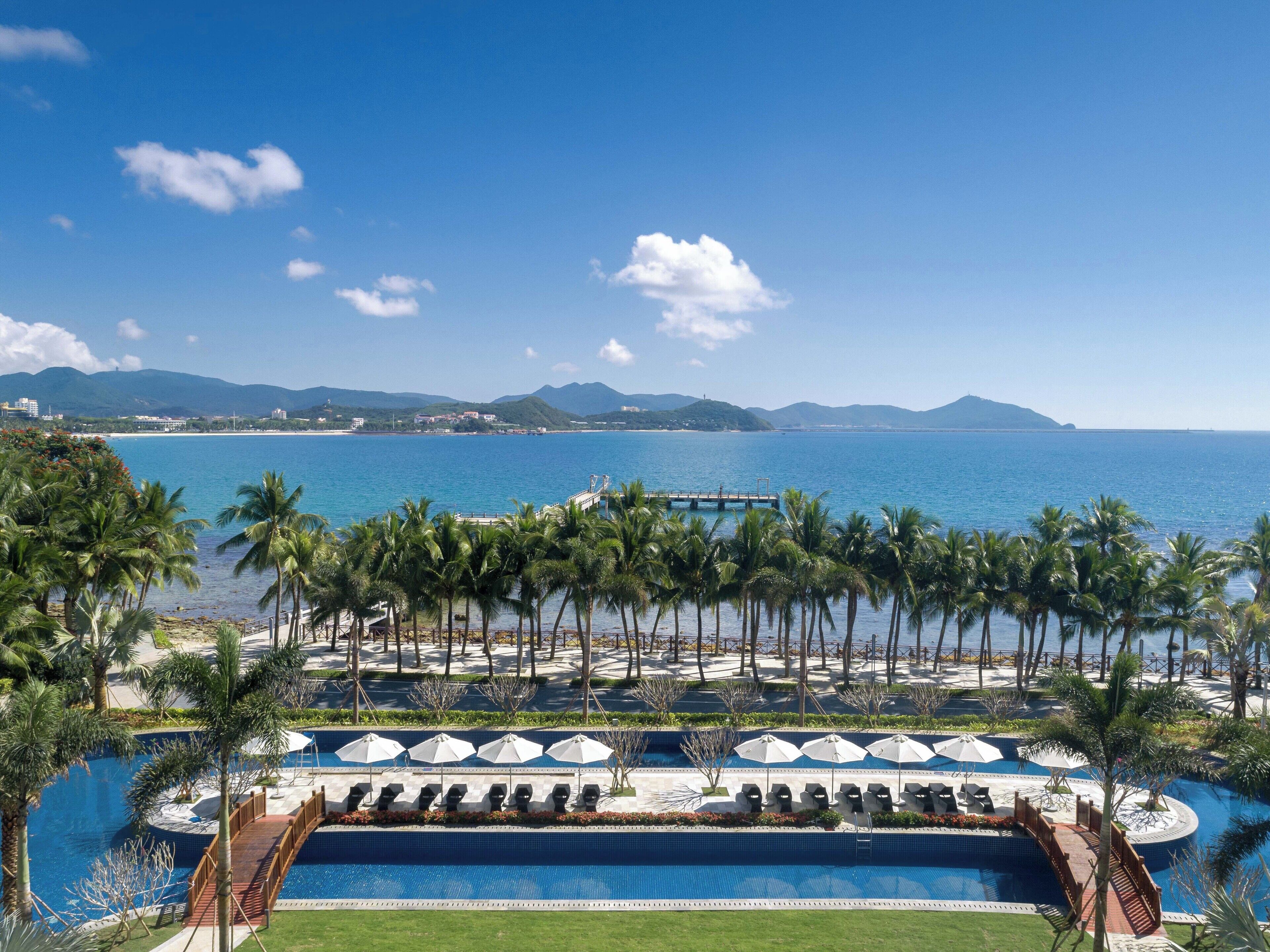 Area piscina di un resort con palme, un ponte e vista sull'oceano e sulle montagne.