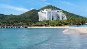 A large, multi-story hotel with a modern design, situated on a beach with a backdrop of lush green hills.