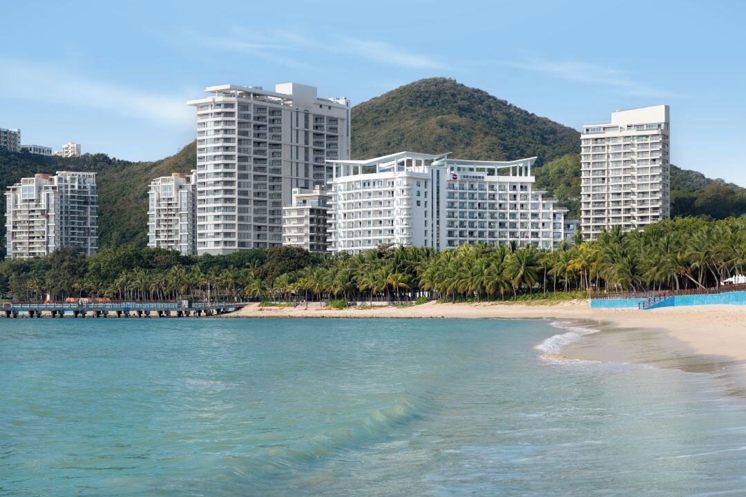 A beachfront hotel with multiple high-rise buildings, a sandy beach, and a forested hill in the background.