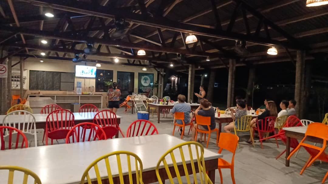 An outdoor dining area with tables and chairs, a stage with a screen, and a wooden ceiling structure.