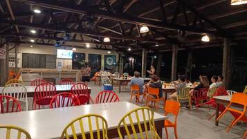 An outdoor dining area with tables and chairs, a stage with a screen, and a wooden ceiling structure.