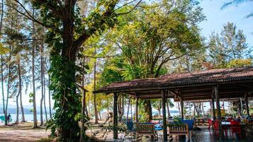 A covered outdoor seating area with tables and chairs, surrounded by trees and a view of the sea.