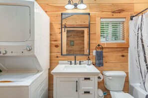 Un baño pequeño con lavadora blanca, lavamanos, inodoro y ventana con persianas.