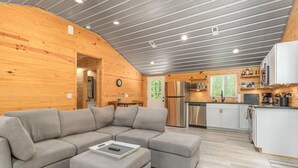 Interior d'una cabana de fusta moderna, amb un sofà modular, una cuina amb electrodomèstics d'acer inoxidable i un sostre de fusta.