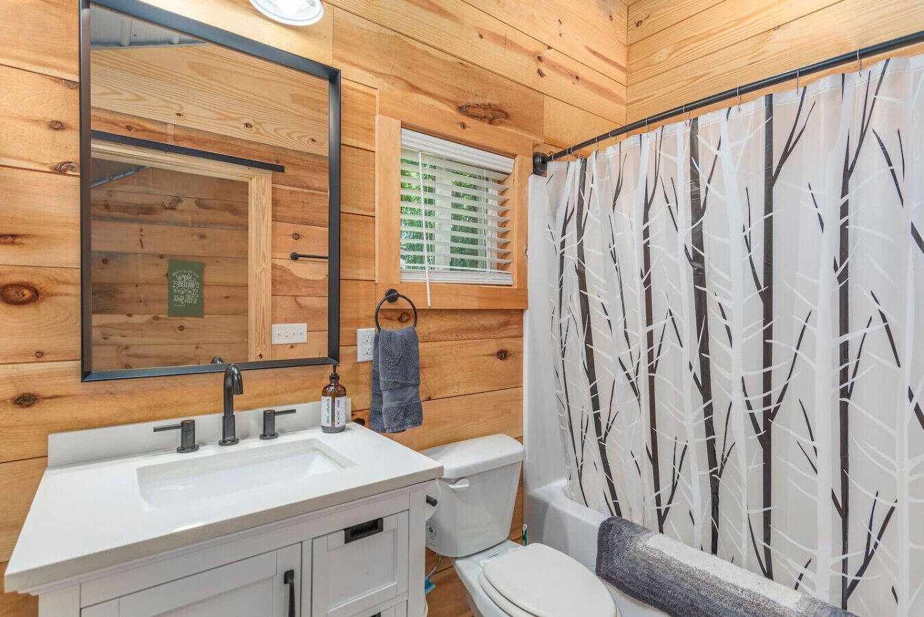 Une salle de bain avec des murs en bois, un lavabo blanc, un miroir, une serviette et un rideau de douche orné d’un motif d’arbre.